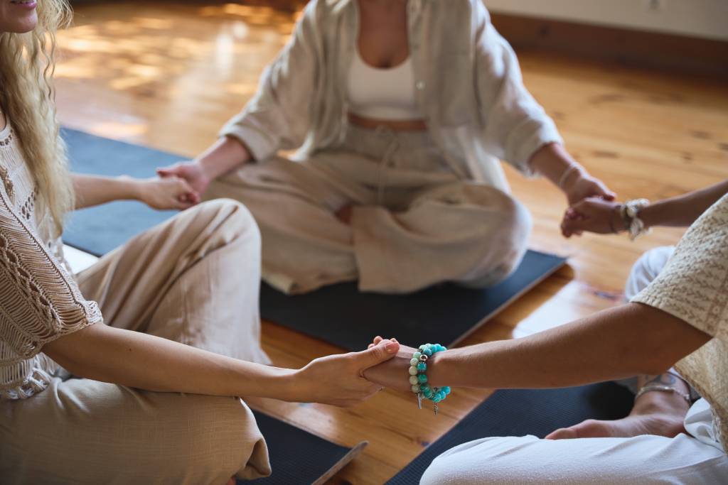 Three diverse women group sitting in circle meditating doing yoga holding hands.